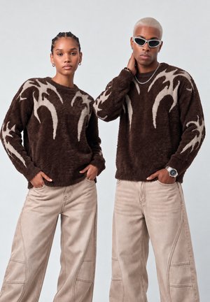 Deux mannequins portant des pulls duveteux brun foncé assortis avec des motifs de flammes beiges et des pantalons amples beige clair sur un fond uni.