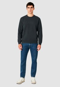 Felpa grigio scuro con logo "GAUDI" in rilievo, scollo tondo, polsini e orlo a costine, abbinata a jeans blu e scarpe da ginnastica bianche.