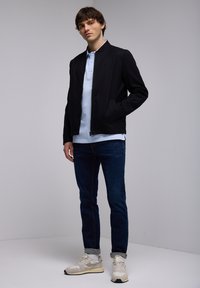 Svart bomberjacka, ljusblå polo, mörka jeans och beige sneakers. Jackan har en framdragkedja och sidofickor.