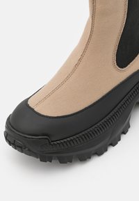 Botte Chelsea en tissu beige et caoutchouc noir avec une semelle épaisse crantée et un panneau latéral élastique, vue de face en angle en gros plan.