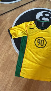 Sárga és zöld sportmez rövid ujjakkal, fekete díszítéssel, és kör alakú logóval, amelyen a "90" szám szerepel. Könnyű anyagból készült.