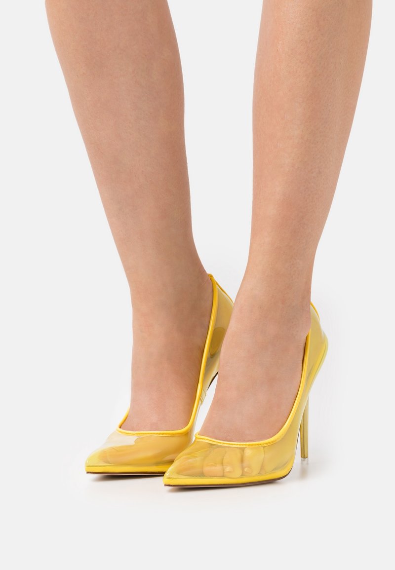 Jambes portant des escarpins transparents jaunes à bouts pointus avec talons stilettos sur fond blanc.