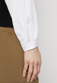 Bílá košilová košile s dlouhými rukávy, se slim fit střihem a manžetami na knoflíky, doplněná o světle hnědé kalhoty. Hladká textura látky.