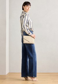 Chemise à manches longues rayée, bleu et blanc ; pochette beige texturée légère ; jean denim à jambes larges ; talons nus à brides ; fond neutre.