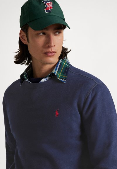 Marinblå sweatshirt med en röd broderad logotyp, lager-på-lager över en rutig skjorta i grönt, blått och vitt. Grön keps med logotypdetaljer.