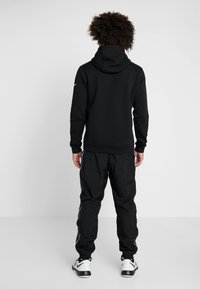 Sweat à capuche noir à coupe décontractée, associé à un pantalon de jogging noir avec des bandes blanches sur les côtés et des poignets élastiques aux chevilles.