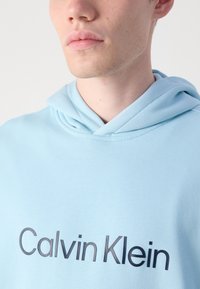 Svetlo modrá mikina s kapucňou s hladkým povrchom a výrazným tmavým logom "Calvin Klein" umiestneným na prednej strane.