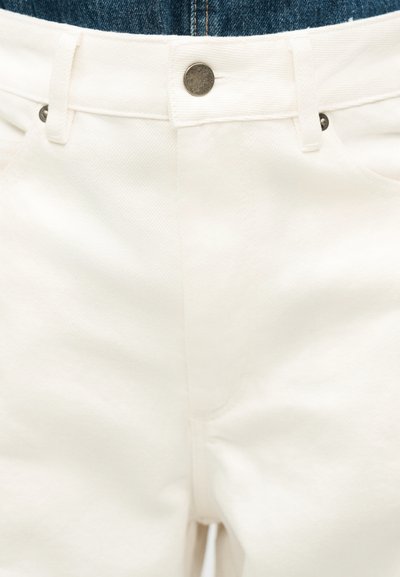 Κοντινή λήψη της περιοχής της μέσης άσπρου τζιν με μεταλλικό κουμπί και θηλιές για ζώνη, φορεμένο πάνω από μπλε τζιν.