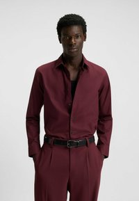 Maroon button-up shirt with black buttons and collar, paired with high-waisted trousers and a black belt. Smooth texture, tailored fit.