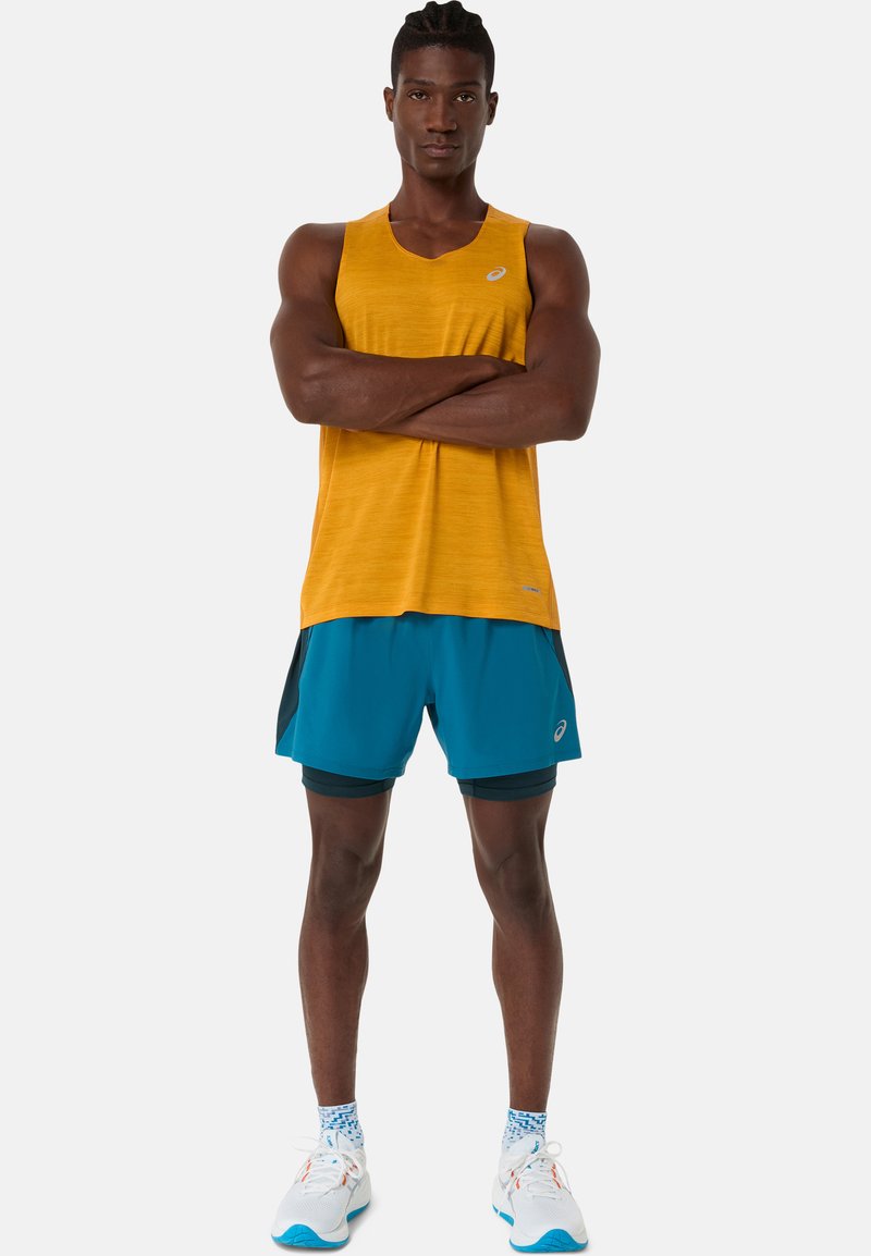 T-shirt de sport jaune à finition texturée, associé à un short turquoise avec des accents noirs. Baskets blanches avec des détails bleus et des chaussettes à motifs.