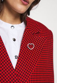 Femme portant un blazer pied-de-poule rouge et noir avec une broche en forme de cœur ornée de strass sur une chemise blanche boutonnée.