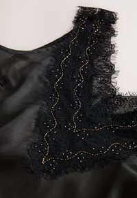 Črna satenasta majica z teksturiranim čipkastim izrezom z bisernimi detajli in raztrganimi robovi, ki poudarjajo zapletene vzorce in kontrastne materiale.