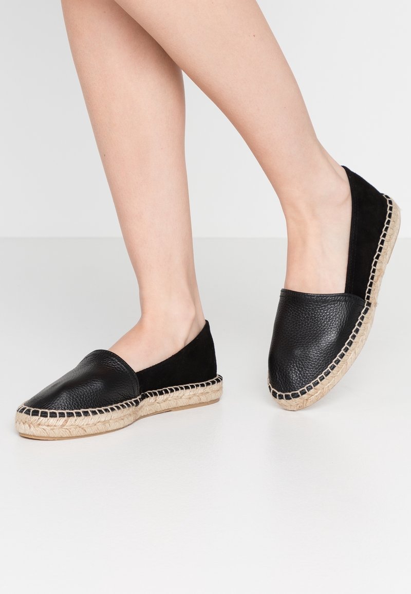 Chaussures espadrilles en cuir noir avec semelles en jute tissée portées par une personne debout sur un sol blanc.