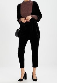 Femme portant un pantalon en velours noir, un cardigan ouvert assorti, un haut à manches longues pailleté marron, des talons noirs et un petit sac à bandoulière noir.