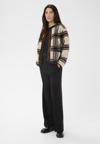 Karierte Fleece-Jacke in Creme, Braun und Blau mit Knopfleiste, kombiniert mit schwarzen Hosen mit weitem Bein und glänzenden schwarzen Schuhen.