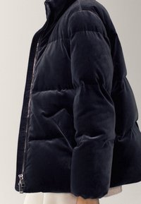 Osoba nosząca ciemnogranatową aksamitną kurtkę puchową z przednim zamkiem błyskawicznym i bocznymi kieszeniami, pokazana od pasa w górę z boku.