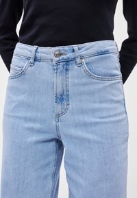 Ανοιχτό μπλε τζιν παντελόνι με ψηλή μέση, σε στυλ πεντασοστού, με κλείσιμο από ασημί κουμπί, πέντε τσέπες και διακριτική φθορά στο μπροστινό ύφασμα.
