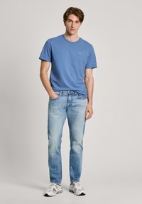 Camiseta azul de cuello redondo combinada con jeans celestes. La camiseta tiene un pequeño detalle de logo; los jeans están ligeramente desgastados. Zapatillas casuales completan el conjunto.