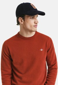 Roter Strickpullover mit Rundhalsausschnitt, mit kleinem Logo auf der linken Seite. Marineblaues Cordhut mit einem "G"-Emblem auf der Vorderseite.