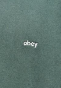 Πράσινο ύφασμα με λευκό κεντημένο πεζό κείμενο "obey" κεντραρισμένο στην επιφάνεια.