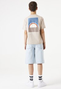 Beige Grafik-T-Shirt mit einem bunten Retro-Design auf der Rückseite, kombiniert mit hellblauen Denim-Shorts und weißen Turnschuhen mit schwarzen Socken.