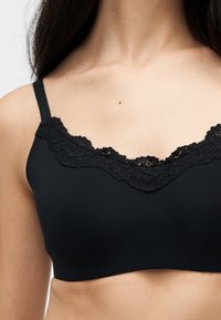 Γυναίκα που φοράει ένα μαύρο bralette με λεπτές τιράντες και δαντέλα κατά μήκος του ντεκολτέ, που αποκαλύπτει το πάνω μέρος του στήθους και την κοιλιά.