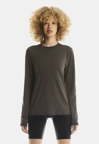 Jong persoon met krullend blond haar, gekleed in een donker, lang mouwshirt en zwarte fietskort, staand tegen een effen witte achtergrond.