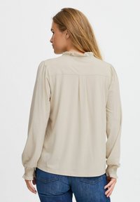 Beige langærmet bluse med smockede manchetter, flæser ved halsen og fold ved ryggen. Glat tekstur og skræddersyet design.