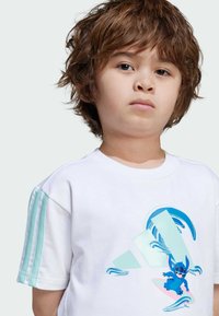 Vit bomullströja med korta ärmar, prydd med mintgröna axelränder och en färgglad grafik av en karaktär som surfar på vågor.