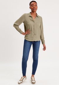 Chemise olive verte à boutons avec un col, deux poches poitrine ; associée à un jean skinny bleu et des baskets beiges et blanches.