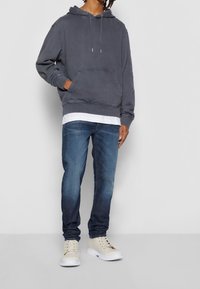 Grå bomullströja med en känguruficka, lagd över en vit skjorta. I kombination med mörkblå jeans och beige höga sneakers.