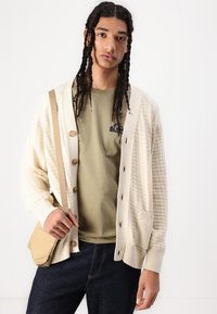 Light cream cardigan with textured pattern, large buttons, and ribbed cuffs. Worn over a green shirt, paired with dark jeans and a beige bag.