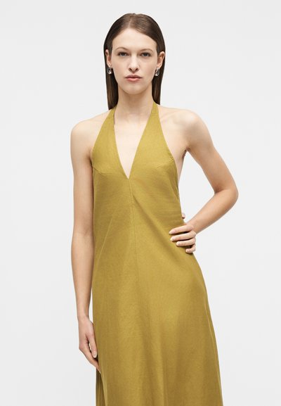 Γυναίκα με ίσια καστανά μαλλιά που φοράει πράσινο φόρεμα με λαιμόκοψη halter, στέκεται με το ένα χέρι στη μέση της μπροστά σε λευκό φόντο.