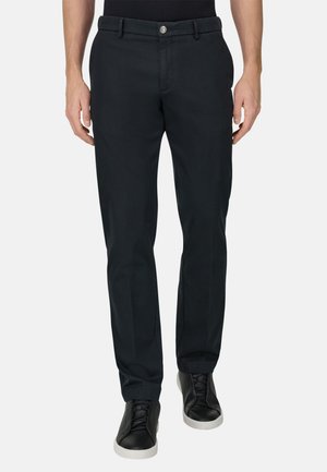 Homme portant un pantalon noir ajusté et des baskets noires à semelles blanches, debout devant un fond clair uni.
