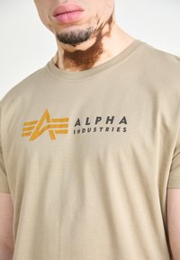 Šviesiai rudos spalvos medvilninis marškinėlis su ryškiu juodu ir oranžiniu "ALPHA INDUSTRIES" logotipu, kuris ant priekinės dalies turi geometrinius dizaino elementus.