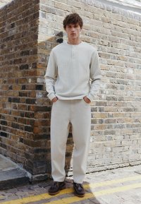 Vaaleanbeige teksturoidulla pitkähihainen paita, kermanväriset väljästi istuvat housut ja tummanruskeat nauhakengät, poseeraten tiiliseinää vasten.