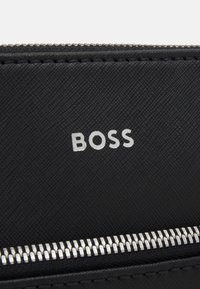 Portefeuille en matériau noir texturé avec des éléments en métal argenté et un logo "BOSS" bien en vue. Détail zip le long du bord inférieur.