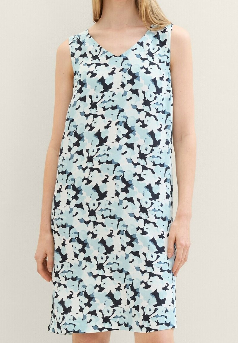 Robe sans manches jusqu'aux genoux avec un col en V et un motif floral abstrait en bleu clair, blanc et noir portée par une personne debout.