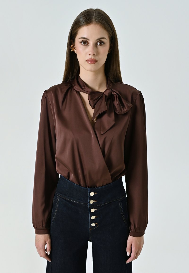 Blusa di raso marrone con un fiocco al collo, maniche lunghe e design avvolgente, abbinata a pantaloni in denim scuro a vita alta con dettagli di bottoni.