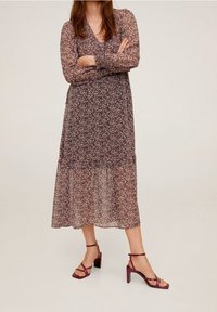Langes, fließendes Kleid aus transparentem Stoff mit einem dunklen Blumenmuster. Das Kleid hat lange Ärmel und einen gestuften Saum. Kombiniert mit bordeauxfarbenen, hochhackigen Sandalen.