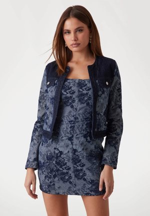 Giacca di denim con trama floreale blu scuro, caratterizzata da un collo rotondo e una silhouette aderente. Abbinata a un mini abito floreale coordinato.