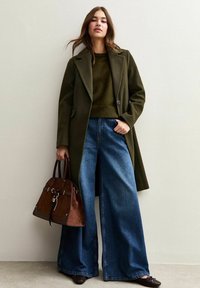 Cappotto verde oliva, maglione verde scuro, jeans a gamba larga blu e borsa marrone con dettagli in metallo; scarpe nere piatte completano l'outfit.
