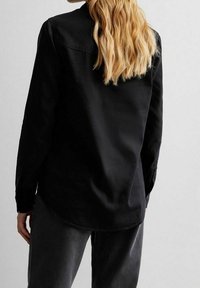 Persona con capelli biondi lunghi indossa una maglietta nera a maniche lunghe e pantaloni scuri, in piedi con le spalle verso di noi su uno sfondo chiaro e uniforme.