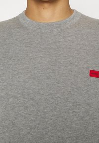 Szary sweter dzianinowy z okrągłym dekoltem. Posiada małą czerwoną metkę z napisem "HUGO" po lewej stronie. Miękka faktura i casualowy design.