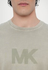 Lysegrå bomulls T-skjorte med et stort, falmet grønt "MK"-logo i sentrum, med klassisk rund hals og subtil tekstur.