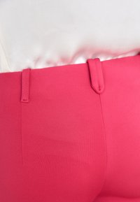 Roze aangesloten broek van gladde, rekbare stof met twee riemlussen in de tailleband. Heeft een effen kleur zonder zichtbare patronen.