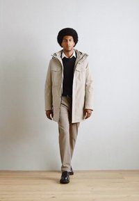 Parka impermeable beige con capucha, dos bolsillos frontales y cierre de cremallera, llevada sobre un suéter negro y pantalones de color crema.