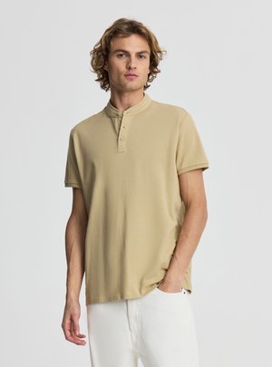 Giovane uomo con capelli ricci che indossa una maglietta henley beige a maniche corte e pantaloni bianchi, in piedi con una mano in tasca.