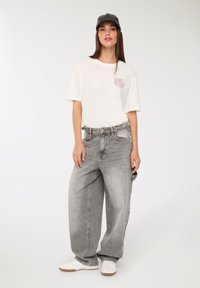 Młoda kobieta w białej koszulce z grafiką, w szerokich, wyblakłych szarych dżinsach, białych sneakersach i czapce baseballowej w kolorze czarnym, stojąca na tle białym.