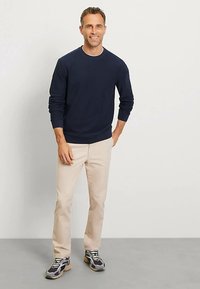 Marineblauwe sweatshirt met lange mouwen, crèmekleurige broek en sneakers met patroon. Model staat voor een neutrale achtergrond en lacht.